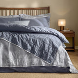 Hebden cotton bedspread in navy and denim blue, shown folded back on a bed to reveal reversible striped and plain percale sides, styled with matching pillows in a wooden-headboard bedroom.