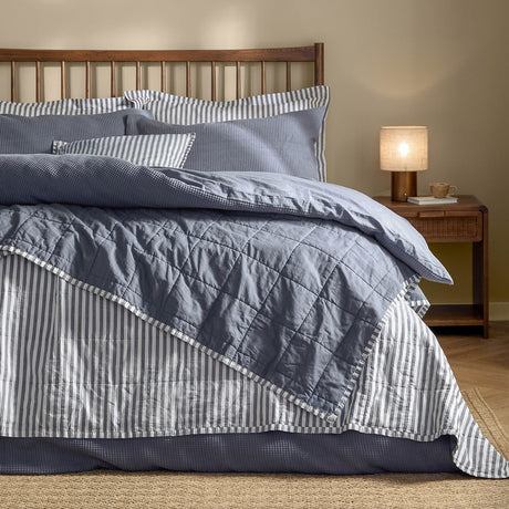 Hebden cotton bedspread in navy and denim blue, shown folded back on a bed to reveal reversible striped and plain percale sides, styled with matching pillows in a wooden-headboard bedroom.