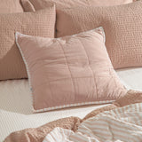 Hebden Percale Cotton Cushion in blush, square quilted design with striped piped edge, styled on a bed with textured pink pillows and striped linens.