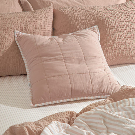 Hebden Percale Cotton Cushion in blush, square quilted design with striped piped edge, styled on a bed with textured pink pillows and striped linens.