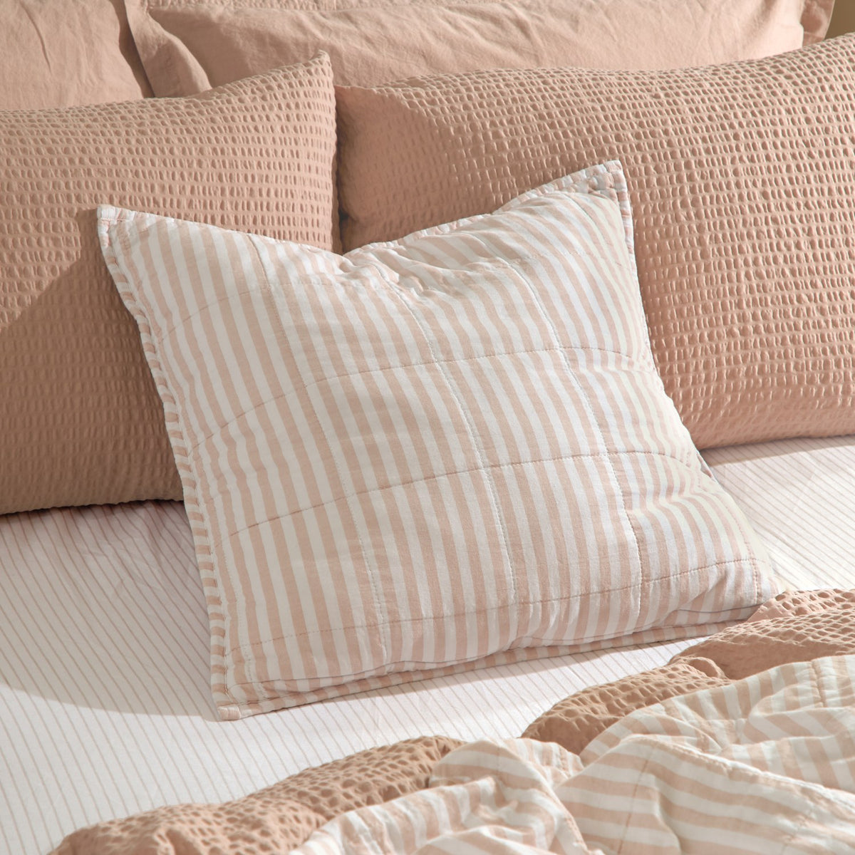 Hebden Percale Cotton Cushion in blush and white stripe with piped edge, styled on a bed against textured blush pillows and a striped duvet.
