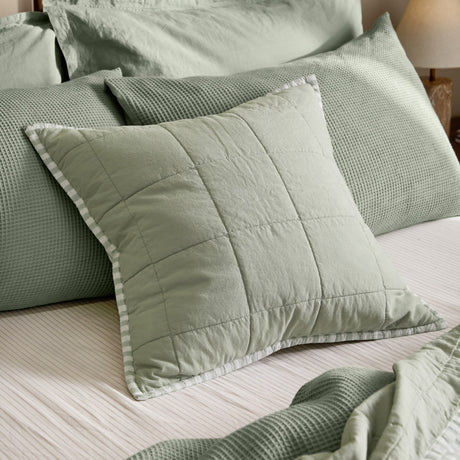 Hebden Percale Cotton Cushion in eucalyptus bay green, square quilted sage cushion with striped edge trim, styled on a bed with waffle-textured pillows.