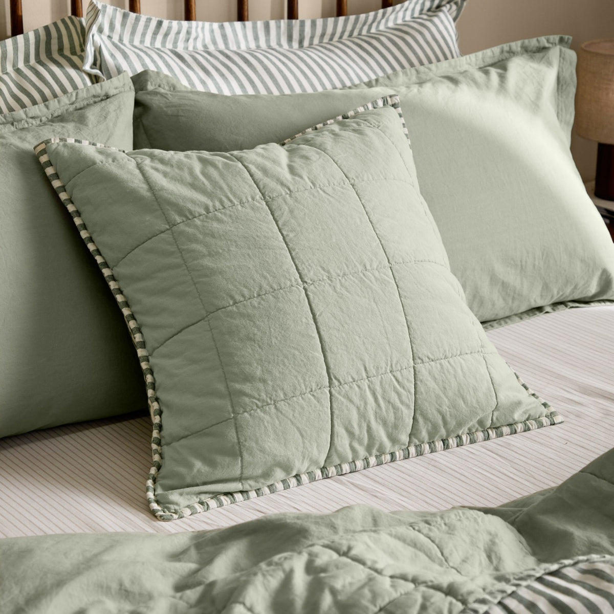 Hebden Percale Cotton Cushion in sage green with quilted grid stitching and striped edge trim, shown among layered pillows on a bed.