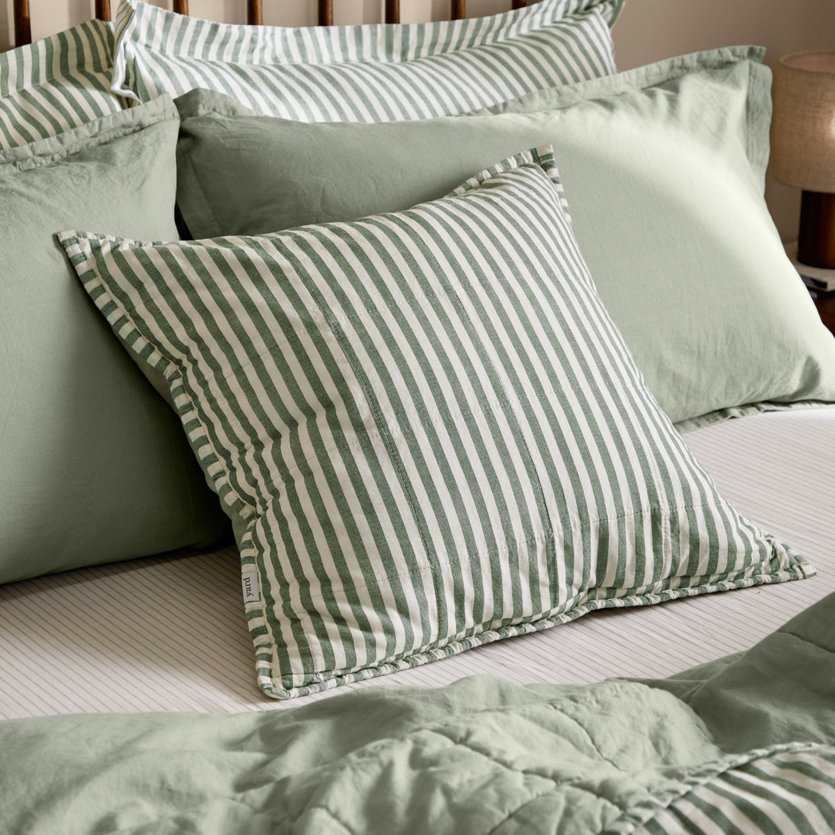 Hebden Percale Cotton Cushion in Forest/Bay Green stripe, shown on a bed layered with sage green pillows, shams, and bedding in a bedroom setting.