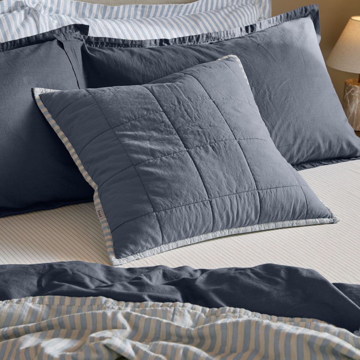 Hebden Percale Cotton Cushion in denim blue with box-quilted grid, shown on a bed layered with striped and navy pillows and bedding.