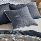 Hebden Percale Cotton Cushion in denim blue with box-quilted grid, shown on a bed layered with striped and navy pillows and bedding.