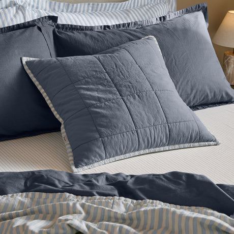 Hebden Percale Cotton Cushion in denim blue with box-quilted grid, shown on a bed layered with striped and navy pillows and bedding.