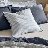 Hebden Percale Cotton Cushion in pale blue and white stripe with gingham-check border edging, styled on a bed alongside denim blue linen pillows and striped bedding.