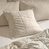 Hebden Percale Cotton Cushion in warm stone with quilted grid stitching and mole grey striped Oxford border, shown on a bed with striped and textured bedding.