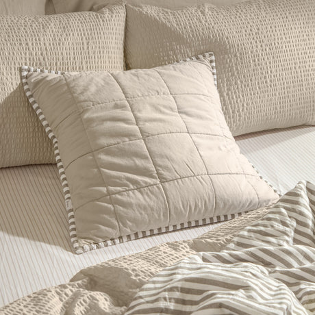 Hebden Percale Cotton Cushion in warm stone with quilted grid stitching and mole grey striped Oxford border, shown on a bed with striped and textured bedding.
