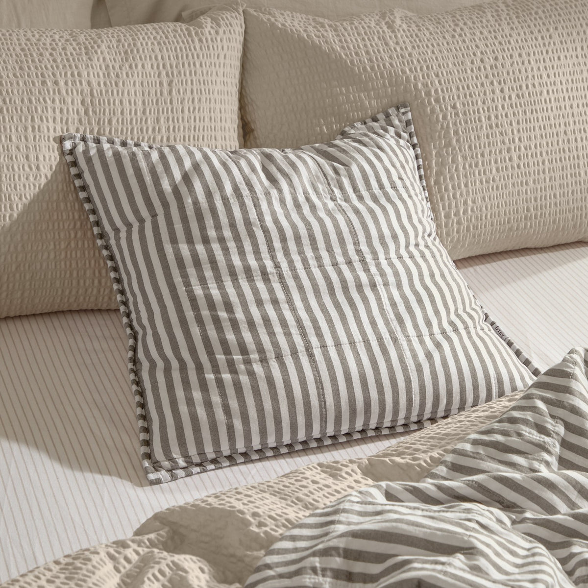 Hebden Percale Cotton Cushion in Mole grey and white stripe with quilted grid and Oxford-style border, shown on a bed with striped sheets and textured cream pillows.