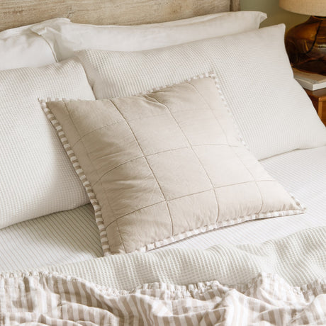 Hebden Percale Cotton Cushion in stone beige with quilted grid front and striped Oxford border, shown on a bed with white waffle pillows.