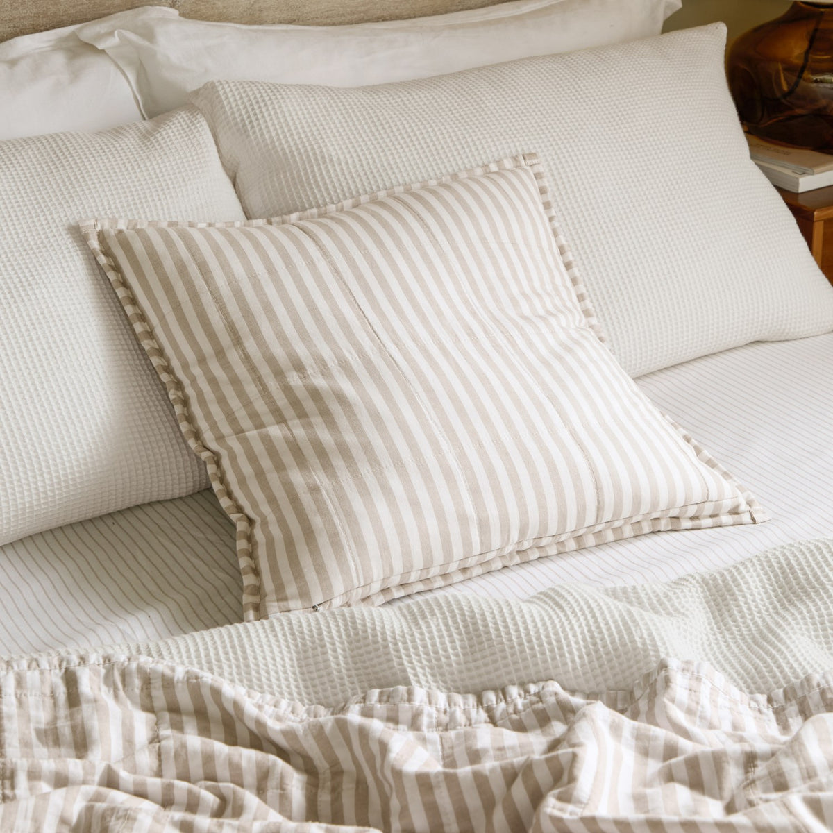 Hebden Percale Cotton Cushion in natural and stone stripe with an Oxford border, styled on a bed against white waffle pillows and striped linen.