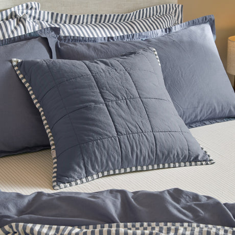 Hebden Percale Cotton Cushion in denim blue with quilted grid stitching and striped navy piping, shown on a bed with matching pillowcases and duvet.
