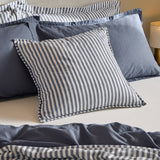 Hebden Percale Cotton Cushion in navy and cream stripe, shown on a bed styled with denim blue linen bedding and pillows.