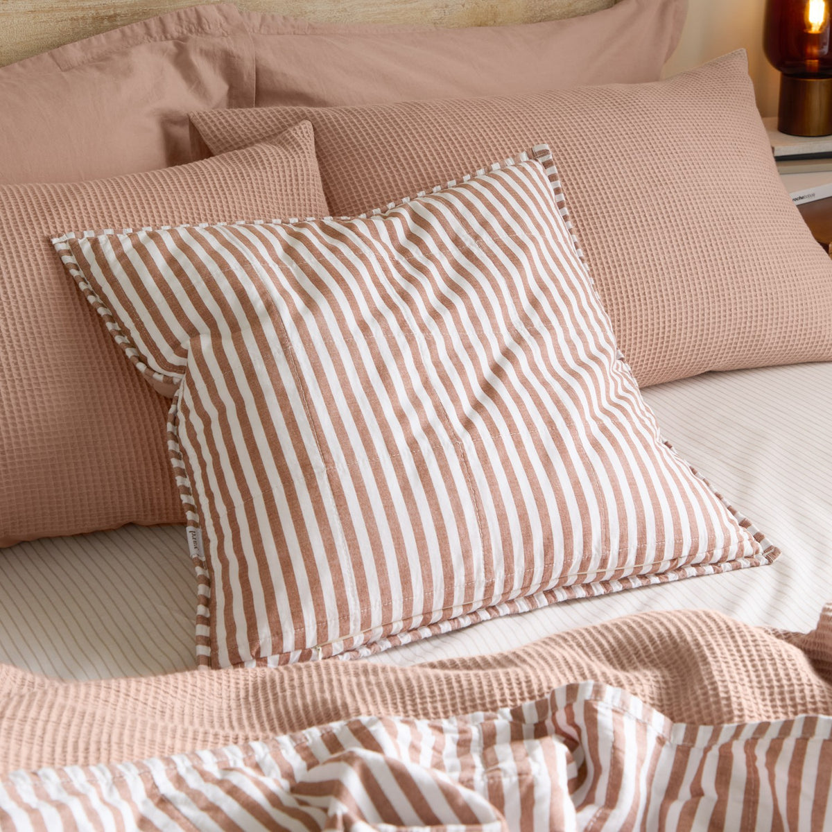 Hebden Percale Cotton Cushion in pecan and white stripe, shown on a bed with pink waffle-textured pillows and matching striped bedding.