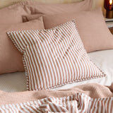 Hebden Percale Cotton Cushion in pecan and white stripe, shown on a bed with pink waffle-textured pillows and matching striped bedding.