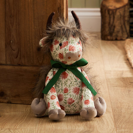 Hugo Polyester Door Stop shaped as a highland cow with shaggy mane, dark horns and grey muzzle, in a coral floral cream fabric with a green ribbon bow, sitting on a wooden floor.