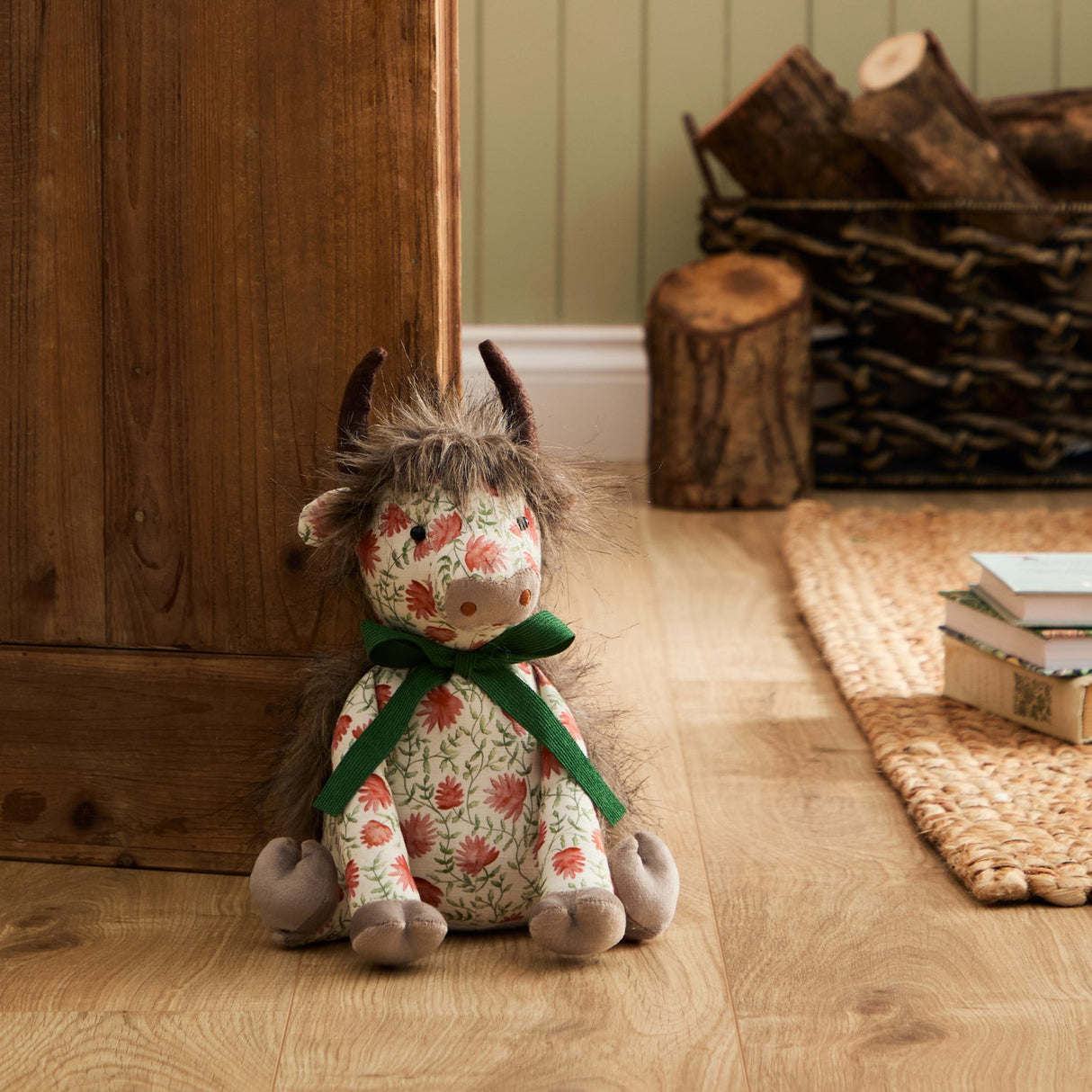 Hugo Polyester Door Stop shaped as a highland cow with shaggy mane, dark horns and grey muzzle, floral cream fabric body tied with a green ribbon bow, shown seated on a wooden floor beside a door.