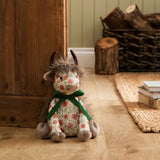 Hugo Polyester Door Stop shaped as a highland cow with shaggy mane, dark horns and grey muzzle, floral cream fabric body tied with a green ribbon bow, shown seated on a wooden floor beside a door.
