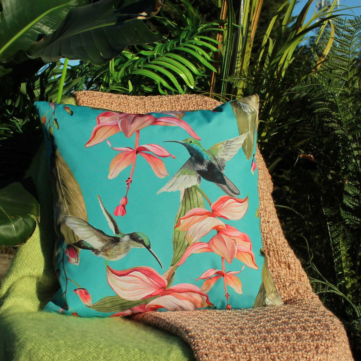 Square polyester cushion in blue-green with two hummingbirds and pink fuchsia flowers printed edge to edge, shown outdoors against tropical foliage.