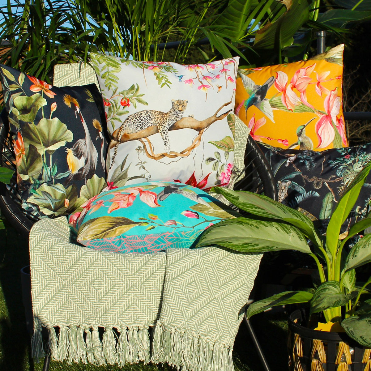 Hummingbird Floral Polyester Cushion in blue, shown centre-front on a garden chair among leopard, crane and floral outdoor cushions with a woven throw.