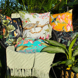 Hummingbird Floral Polyester Cushion in blue, shown centre-front on a garden chair among leopard, crane and floral outdoor cushions with a woven throw.
