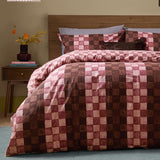 Huxley Reversible Check Duvet Cover Set in Chocolate & Blush, shown on a bed with striped pillowcases and a dark cushion in a bedroom setting with a wooden headboard and side table.