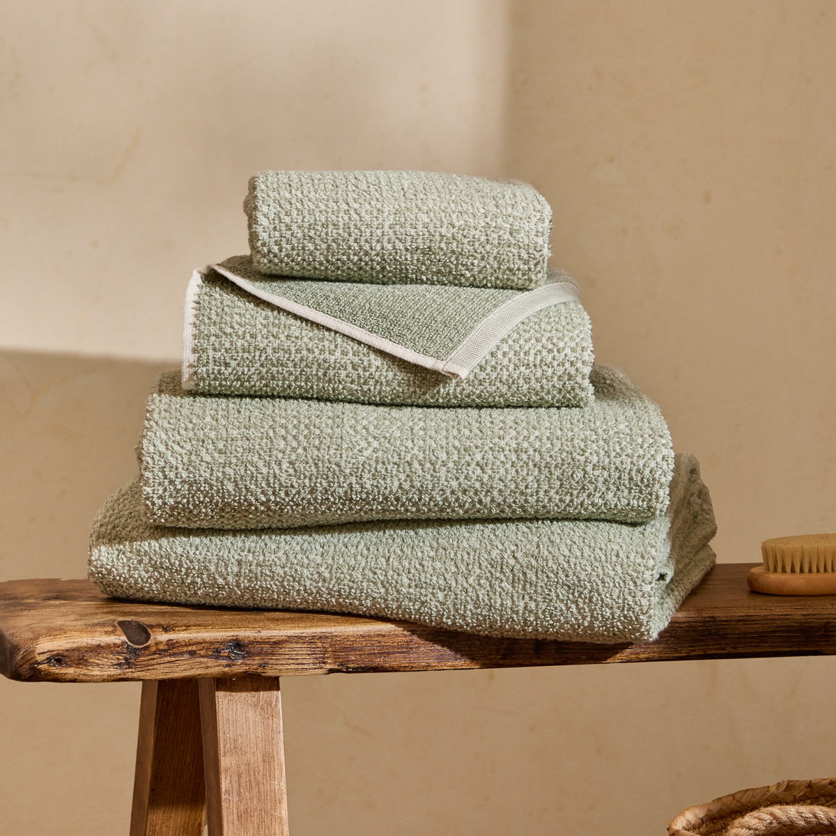 Hebden Plain Cotton Towels in Bay Green, folded stack of four textured waffle-weave towels with off-white contrast border, shown on a wooden bench.