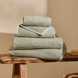Hebden Plain Cotton Towels in Bay Green, folded stack of four textured waffle-weave towels with off-white contrast border, shown on a wooden bench.