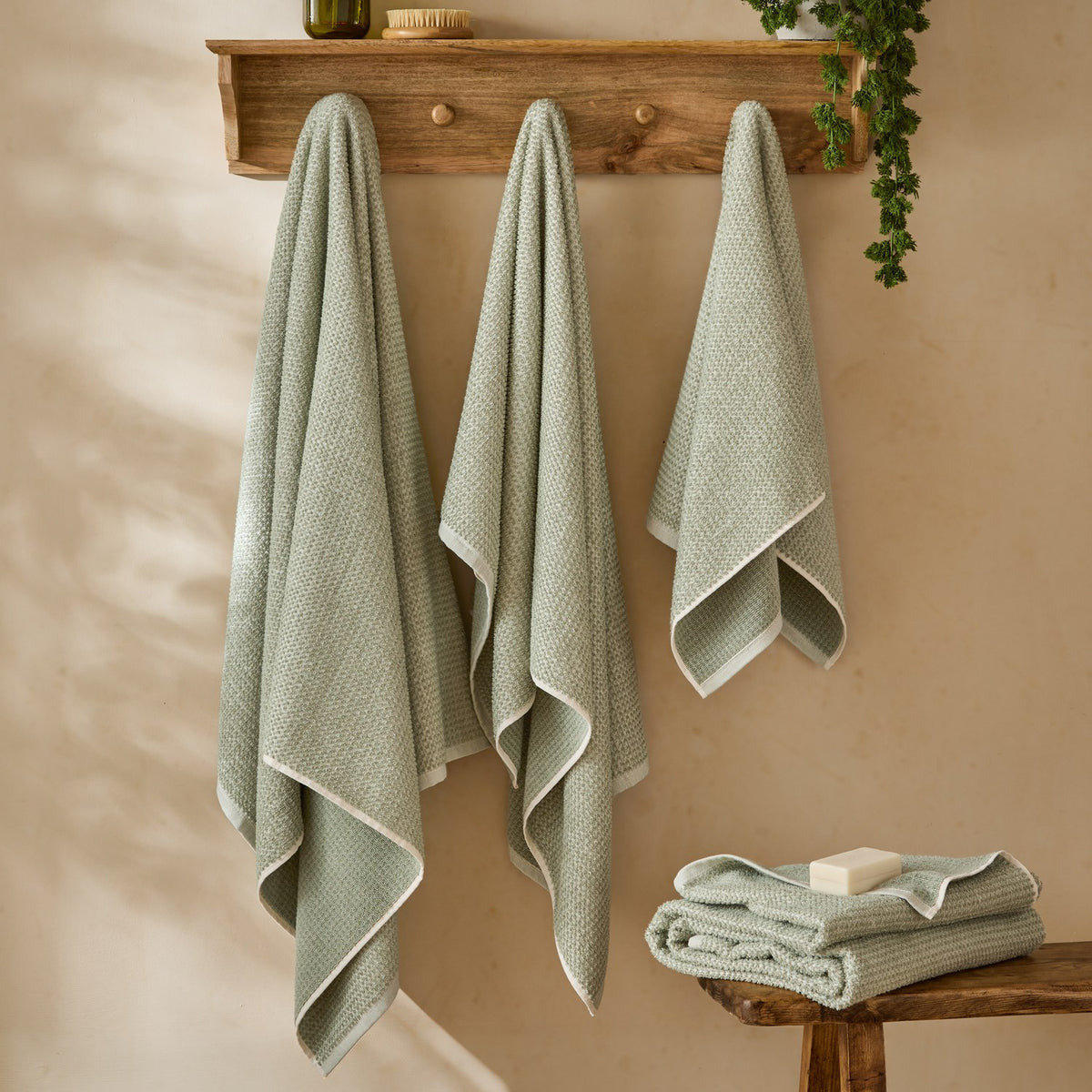Three Hebden waffle-weave cotton towels in bay green with off-white borders hang from wooden wall hooks, with a folded towel and soap on a wooden bench below.