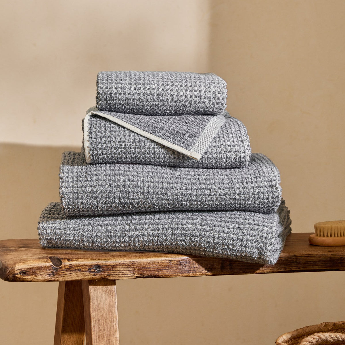 Hebden Plain Cotton Towels in denim blue, four folded waffle-weave towels stacked on a wooden bench with a cream wall background.