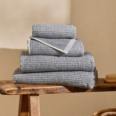 Hebden Plain Cotton Towels in denim blue, four folded waffle-weave towels stacked on a wooden bench with a cream wall background.