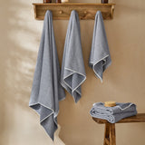 Hebden Plain Cotton Towels in denim blue, three waffle-weave towels with white-edge trim hanging on wooden wall hooks, plus a folded set on a wooden stool in a bathroom setting.