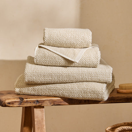 Hebden Plain Cotton Towels in Stone, four folded textured waffle-weave towels stacked on a wooden bench against a neutral background.