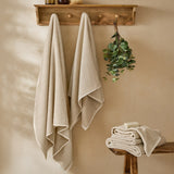 Hebden Plain Cotton Towels in stone, waffle-weave cotton, two hung on wooden wall hooks and folded stack on bench in bathroom setting.