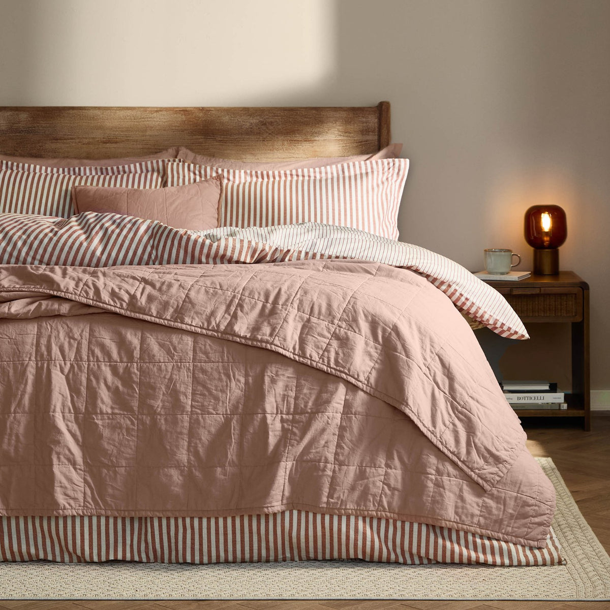 Hebden quilted cotton percale bedspread in pink (Cotta) on a bed with striped pillowcases, wooden headboard, and bedside table in a bedroom setting.