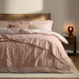 Hebden quilted cotton percale bedspread in pink (Cotta) on a bed with striped pillowcases, wooden headboard, and bedside table in a bedroom setting.