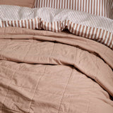 Hebden Percale Cotton Bedspread in Cotta pink, close-up of quilted grid-stitched cotton with striped pillowcases visible behind on the bed.