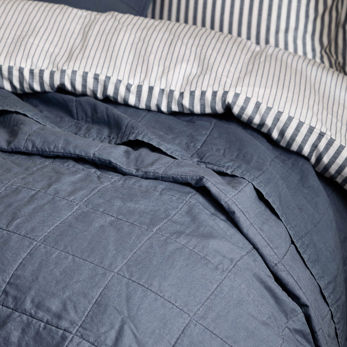 Hebden Percale Cotton Bedspread in denim blue with stitched grid quilting, shown folded back over striped bedding to reveal the matching underside.