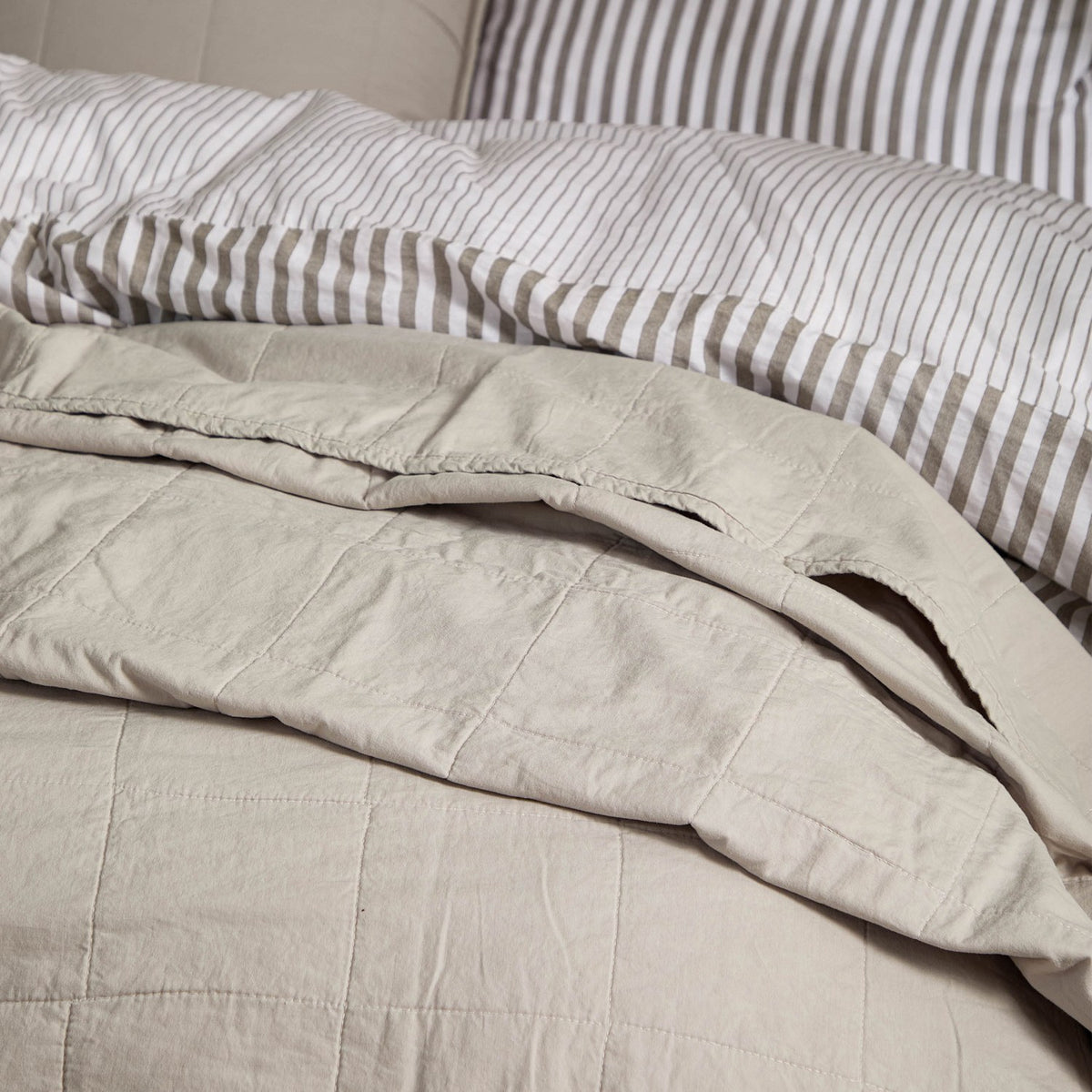 Hebden Percale Cotton Bedspread in stone, softly quilted cotton with a garment-washed, relaxed texture, shown folded on a bed with striped bedding behind.