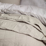 Hebden Percale Cotton Bedspread in stone, softly quilted cotton with a garment-washed, relaxed texture, shown folded on a bed with striped bedding behind.