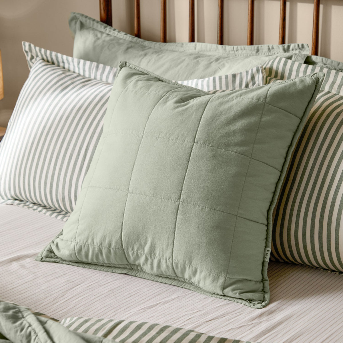 Hebden Percale Cotton Cushion in Bay Green, a quilted grid-stitched square cushion in soft sage cotton, shown on a bed with striped pillowcases.