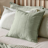 Hebden Percale Cotton Cushion in Bay Green, a quilted grid-stitched square cushion in soft sage cotton, shown on a bed with striped pillowcases.