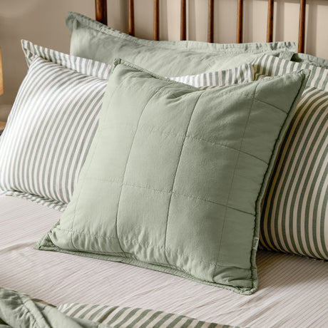 Hebden Percale Cotton Cushion in Bay Green, a quilted grid-stitched square cushion in soft sage cotton, shown on a bed with striped pillowcases.
