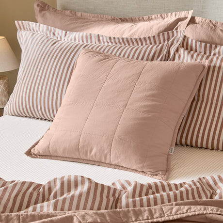 Hebden Percale Cotton Cushion in Cotta pink, quilted into a soft grid with an Oxford border, styled on a bed with striped pillowcases and bedding.