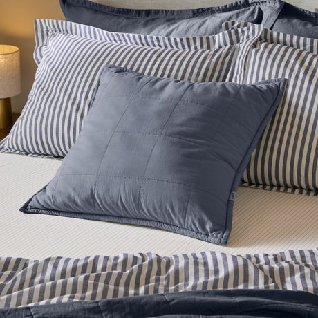 Hebden Percale Cotton Cushion in denim blue with square-grid quilting and Oxford border edge, styled on a bed with striped pillowcases.