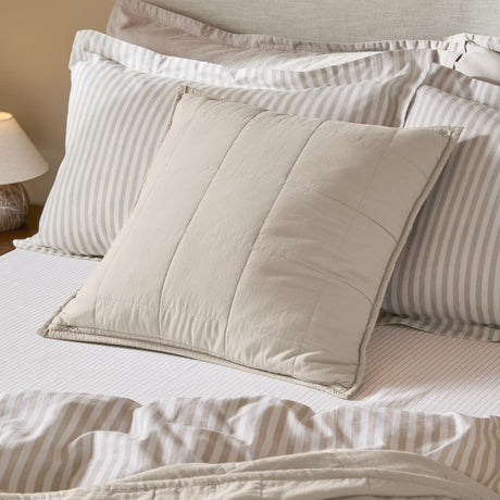 Hebden Percale Cotton Cushion in stone with quilted grid stitching, shown on a bed layered with striped pillowcases and neutral bedding.