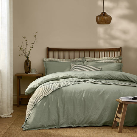 Hebden Cotton Plain Duvet Cover Set in Bay Green on a bed with a striped reverse and pillowcases, shown in a bedroom with a dark wooden headboard.