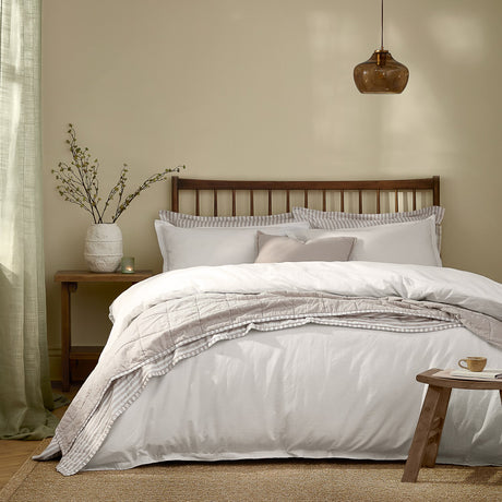 Hebden Cotton Plain Duvet Cover Set in chalk white on a bed with striped pillowcases, quilted throw, dark wood headboard, and bedside table in a bedroom setting.